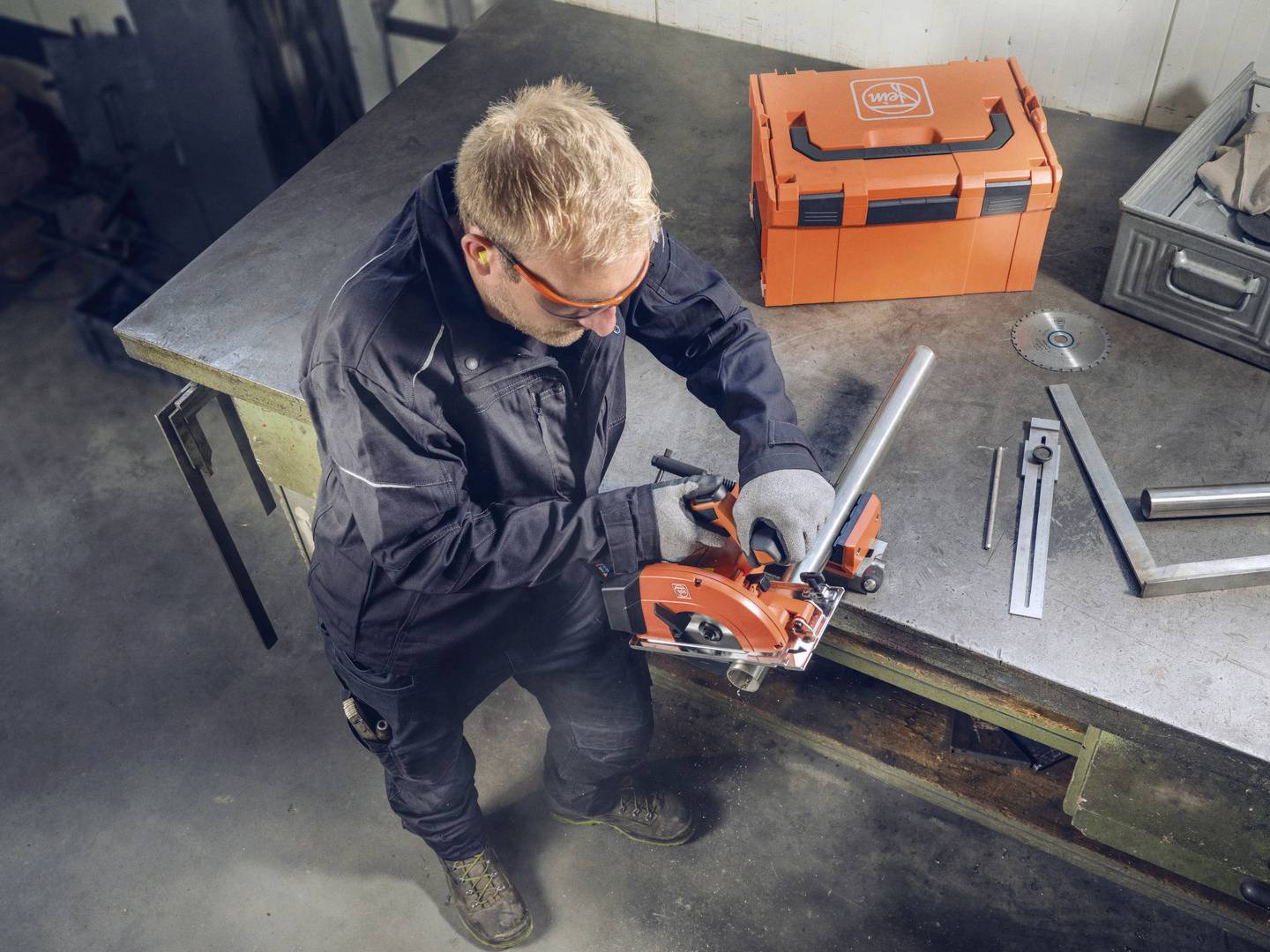 Eine Person in Arbeitskleidung schneidet ein Metallrohr mit einer tragbaren Säge auf einer Werkbank neben einem orangefarbenen Werkzeugkasten.