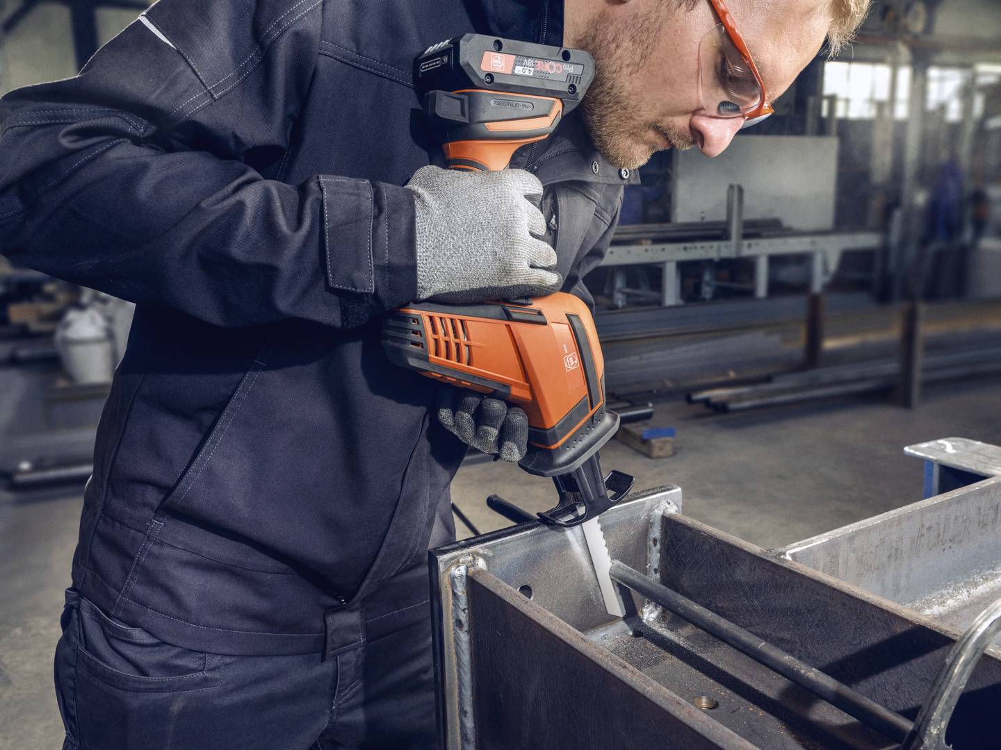Ein Arbeiter in Schutzkleidung schneidet Metall mit einer elektrischen Säge in einer Werkstatt. Im Hintergrund sind Industriegeräte.