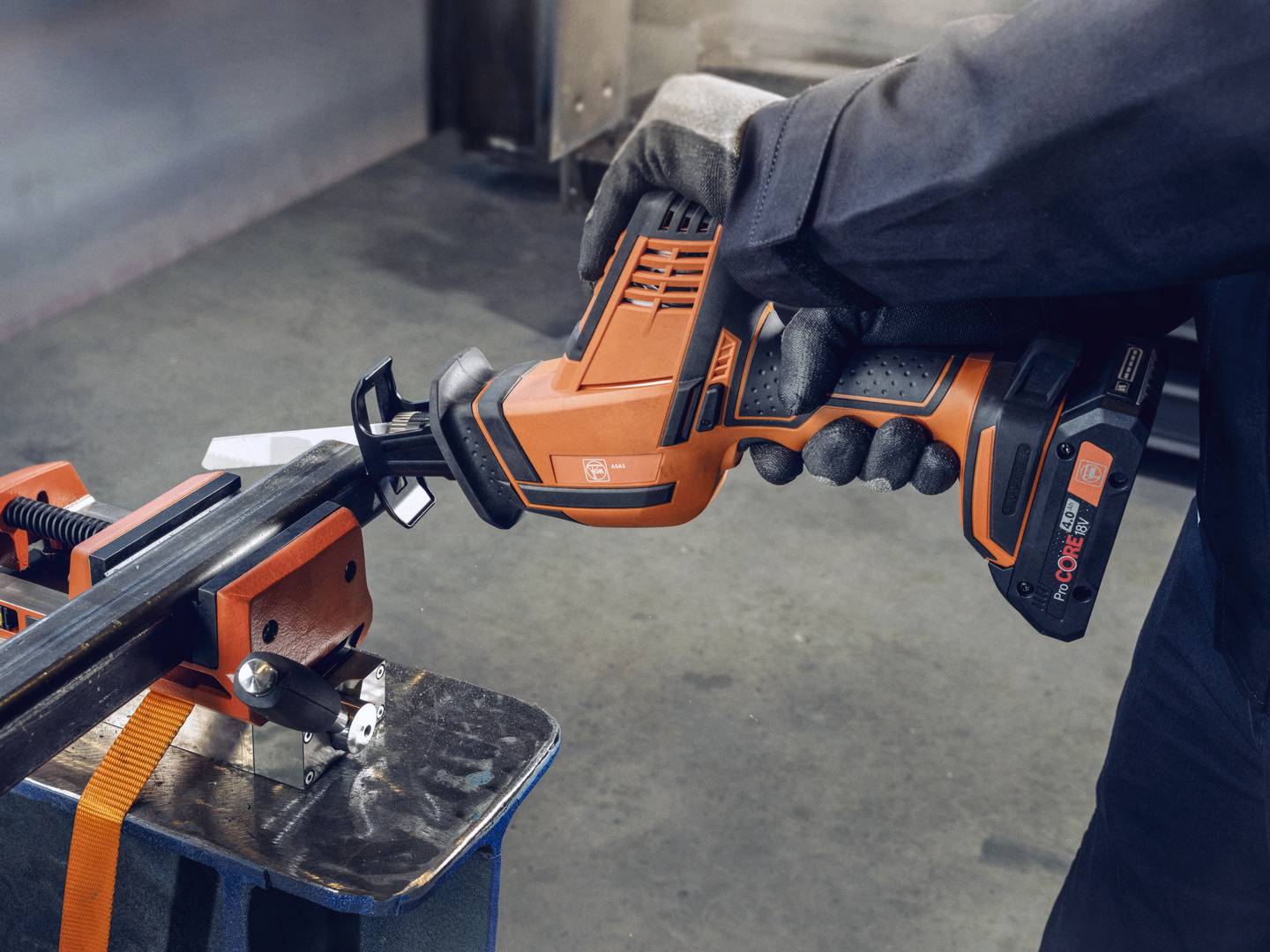 Eine Person benutzt eine orangefarbene, elektrische Säge, um ein Metallrohr in einer Werkstattumgebung zu schneiden.