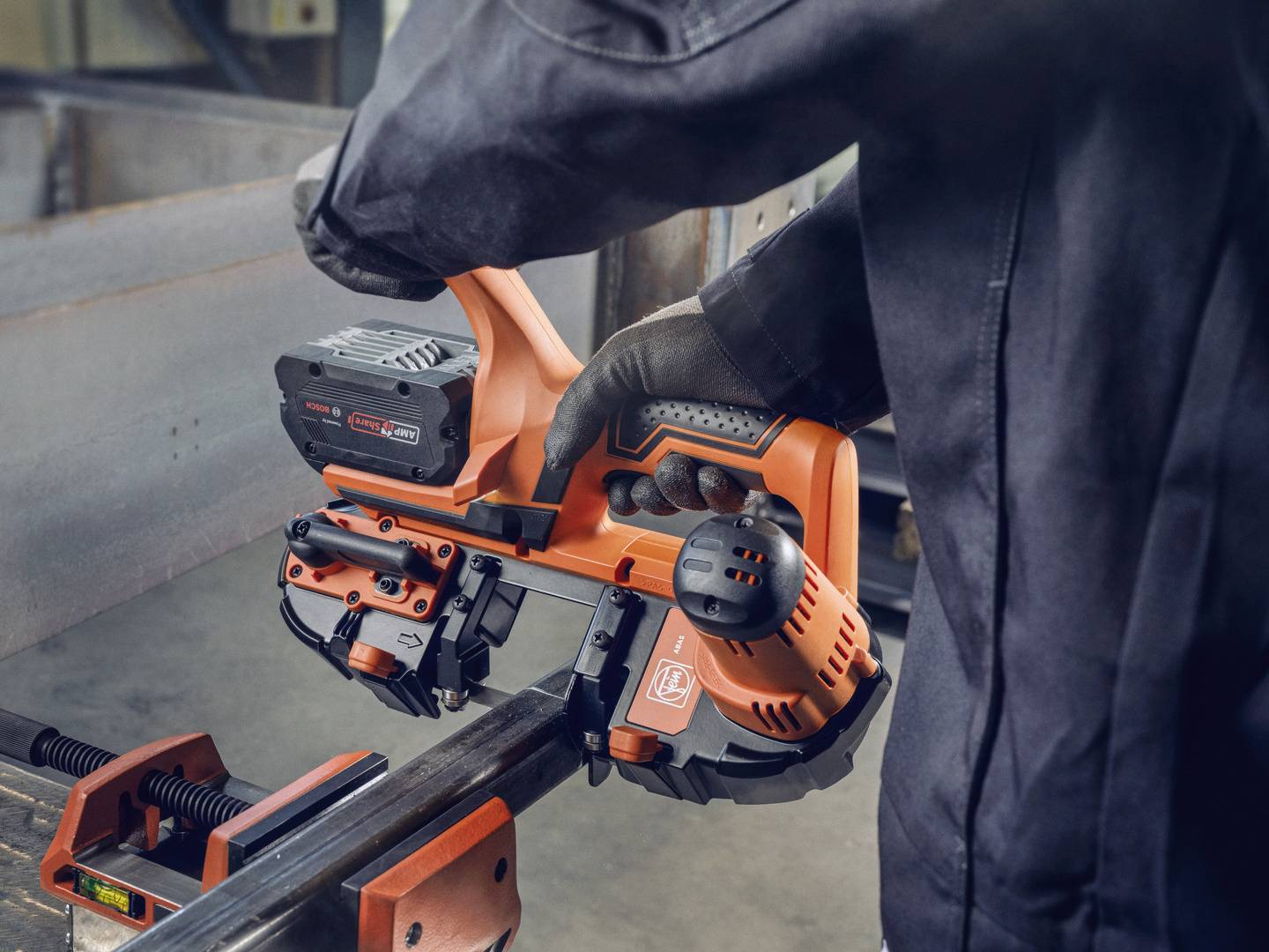 Eine Person in Arbeitskleidung benutzt ein elektrisches Handgerät, um ein Metallstück zu bearbeiten. Der Fokus liegt auf dem Werkzeug.