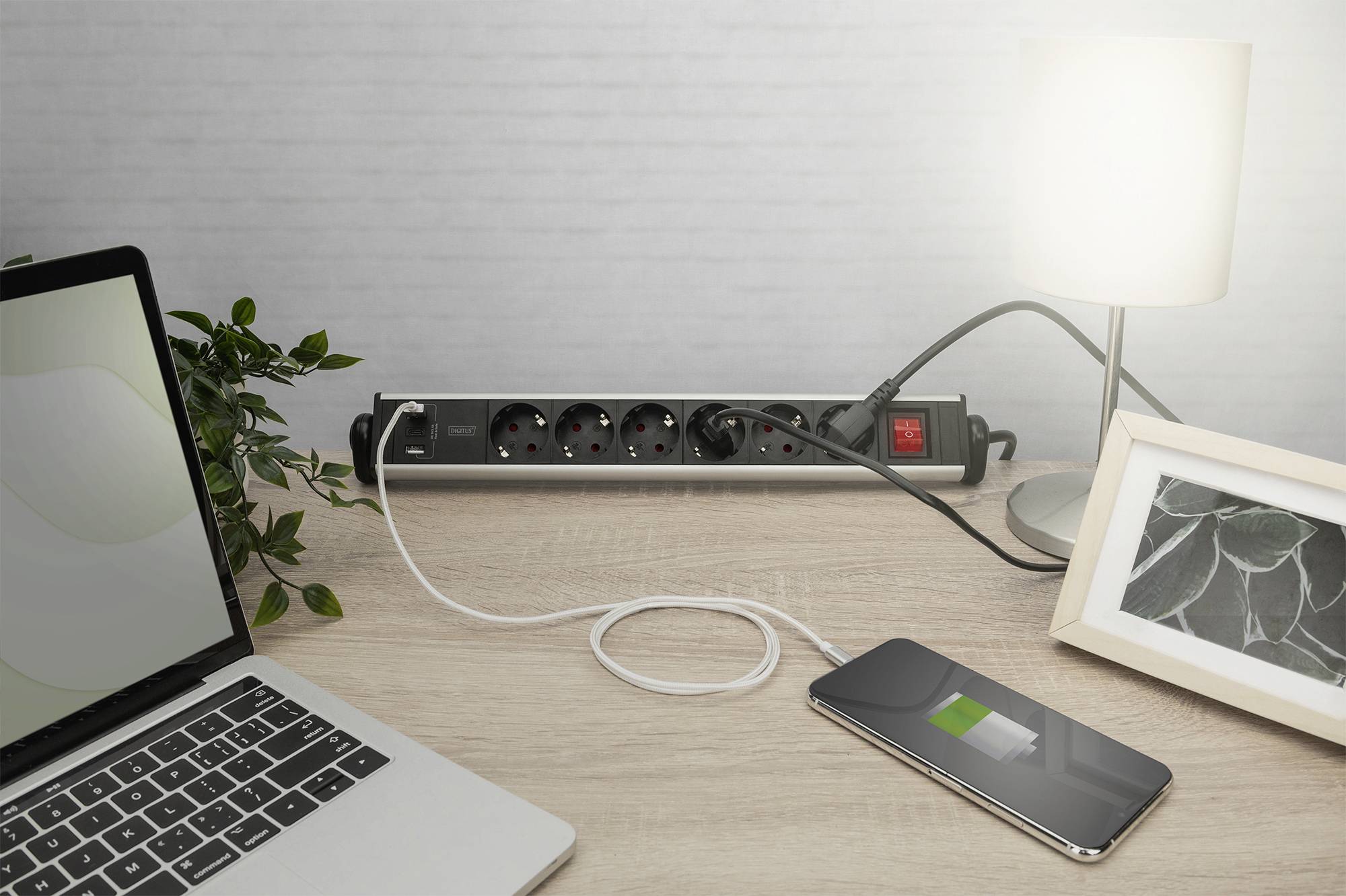 Ein Schreibtisch mit einem Laptop, einem Ladegerät, das ein Smartphone lädt, einer Tischlampe und einem Pflanzenbildrahmen.