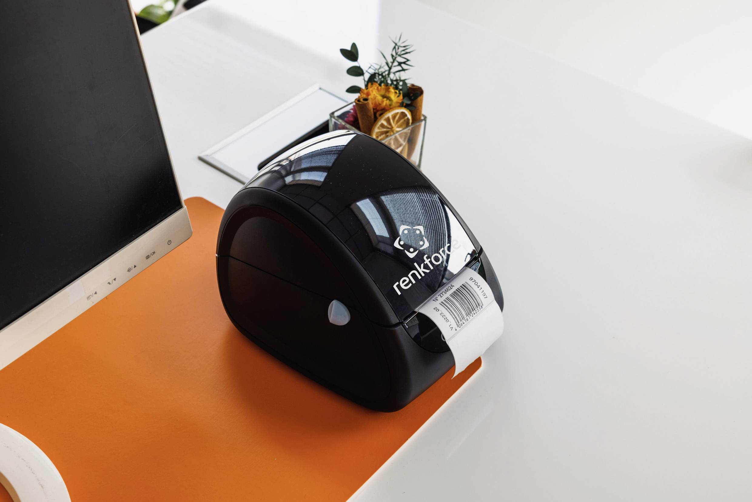 Etikettendrucker auf einem Schreibtisch, druckt ein weißes Etikett. Im Hintergrund sind ein Monitor und eine kleine Pflanze.