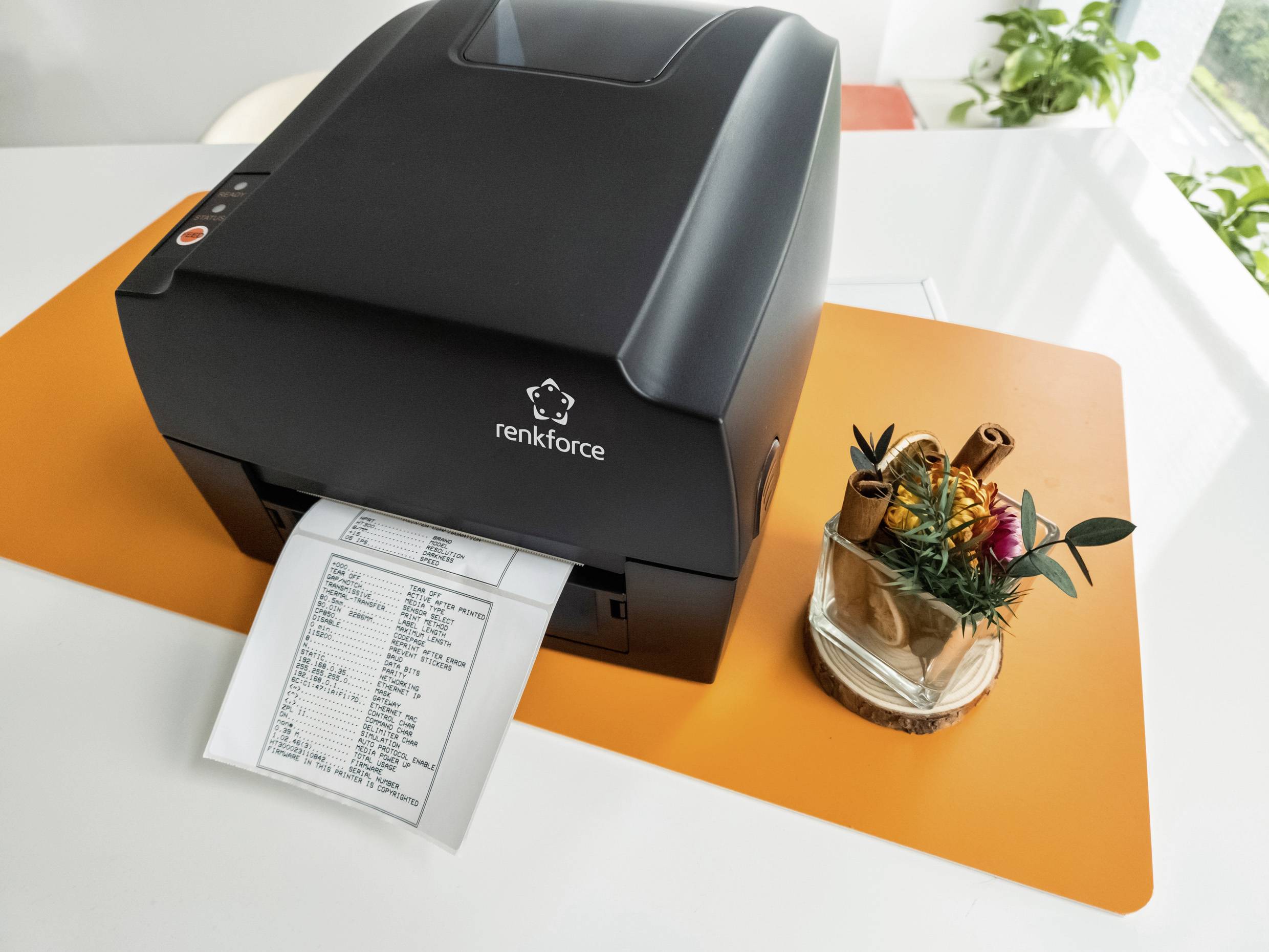 Ein Thermodrucker auf einem Tisch druckt eine Quittung aus. Daneben stehen Pflanzen auf einem orangefarbenen Tischset.