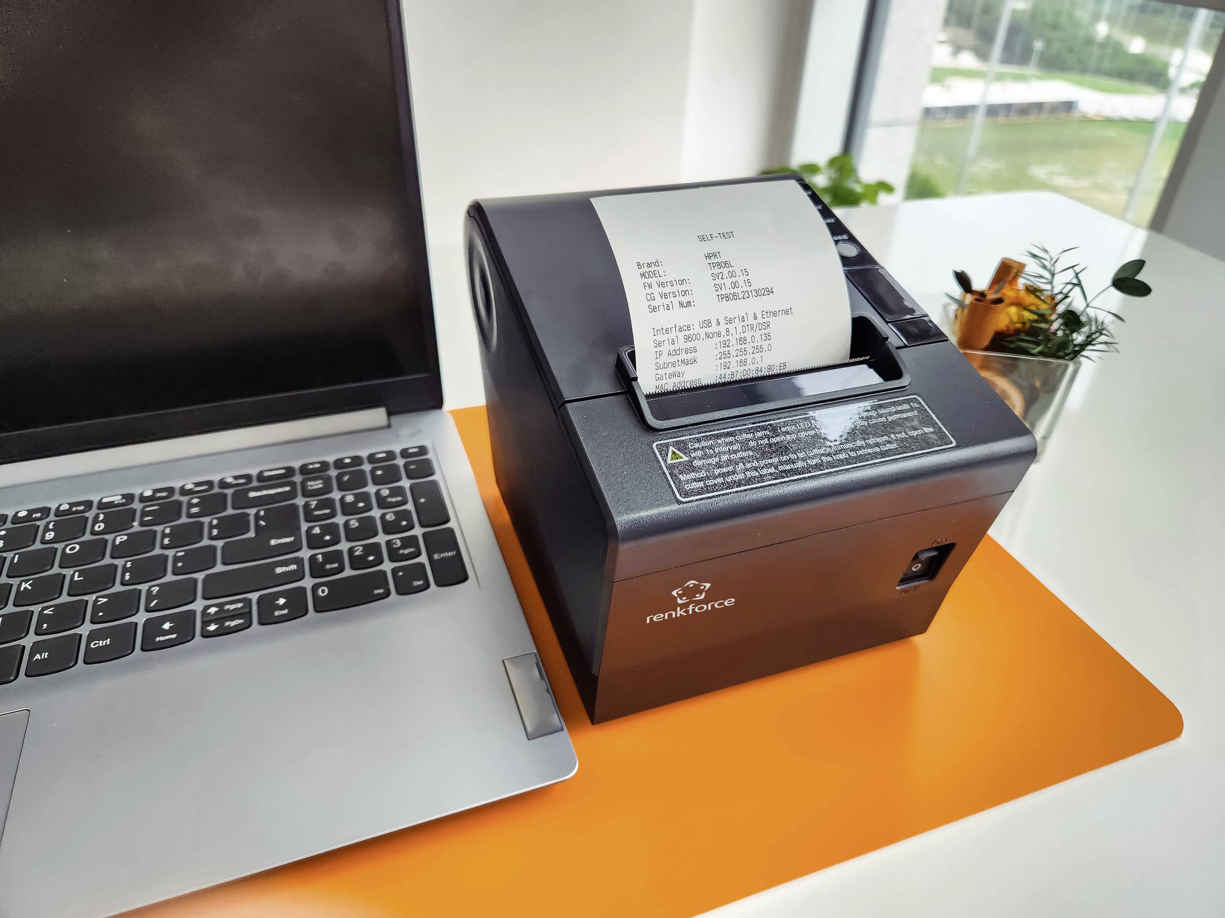 Ein Laptop neben einem Drucker, der einen Kassenbeleg ausgibt. Der Drucker steht auf einem orangefarbenen Tisch, daneben Pflanzen.