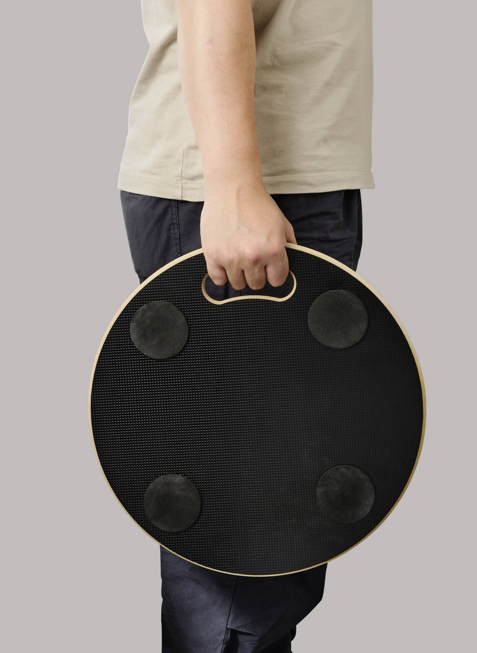 Person hält ein rundes, schwarzes Balance-Board mit Griff und vier rutschfesten Pads in der Hand.
