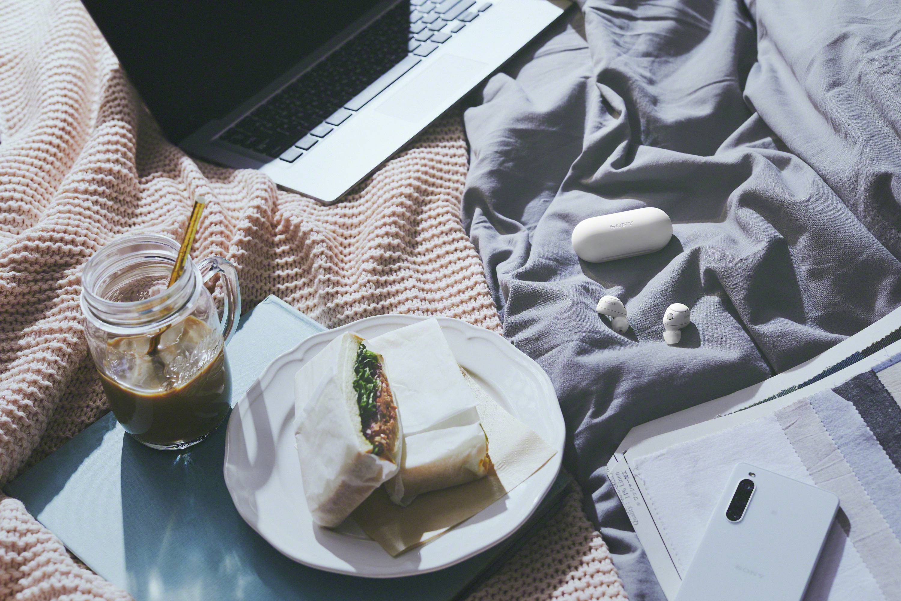 Auf einem Bett liegen ein Laptop, ein Teller mit einem Sandwich, ein Becher Eiskaffee, kabellose Ohrhörer und ein Smartphone auf einem Notizbuch.