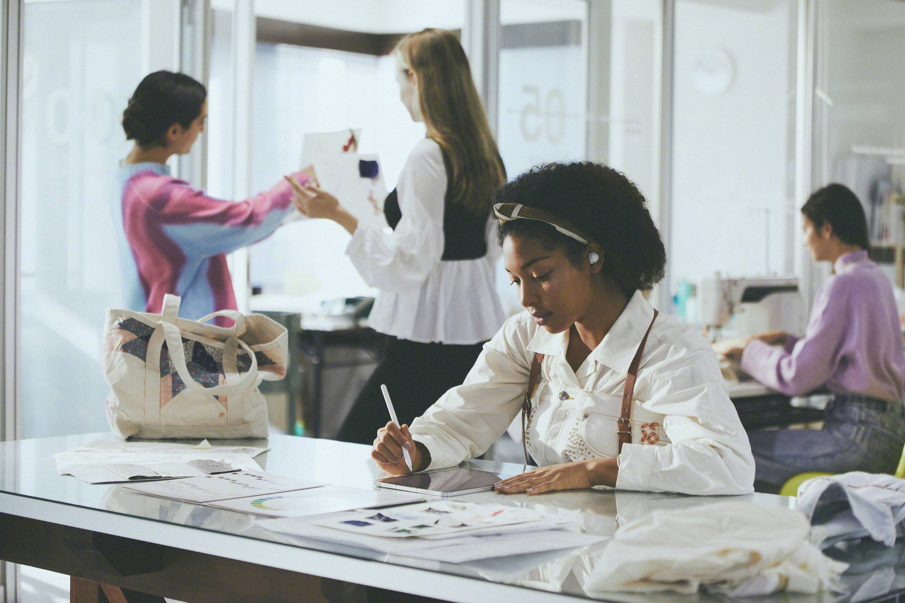 Eine Gruppe von Frauen arbeitet in einem modernen Büro. Im Vordergrund zeichnet eine Frau an einem Schreibtisch, während andere im Hintergrund diskutieren und arbeiten.