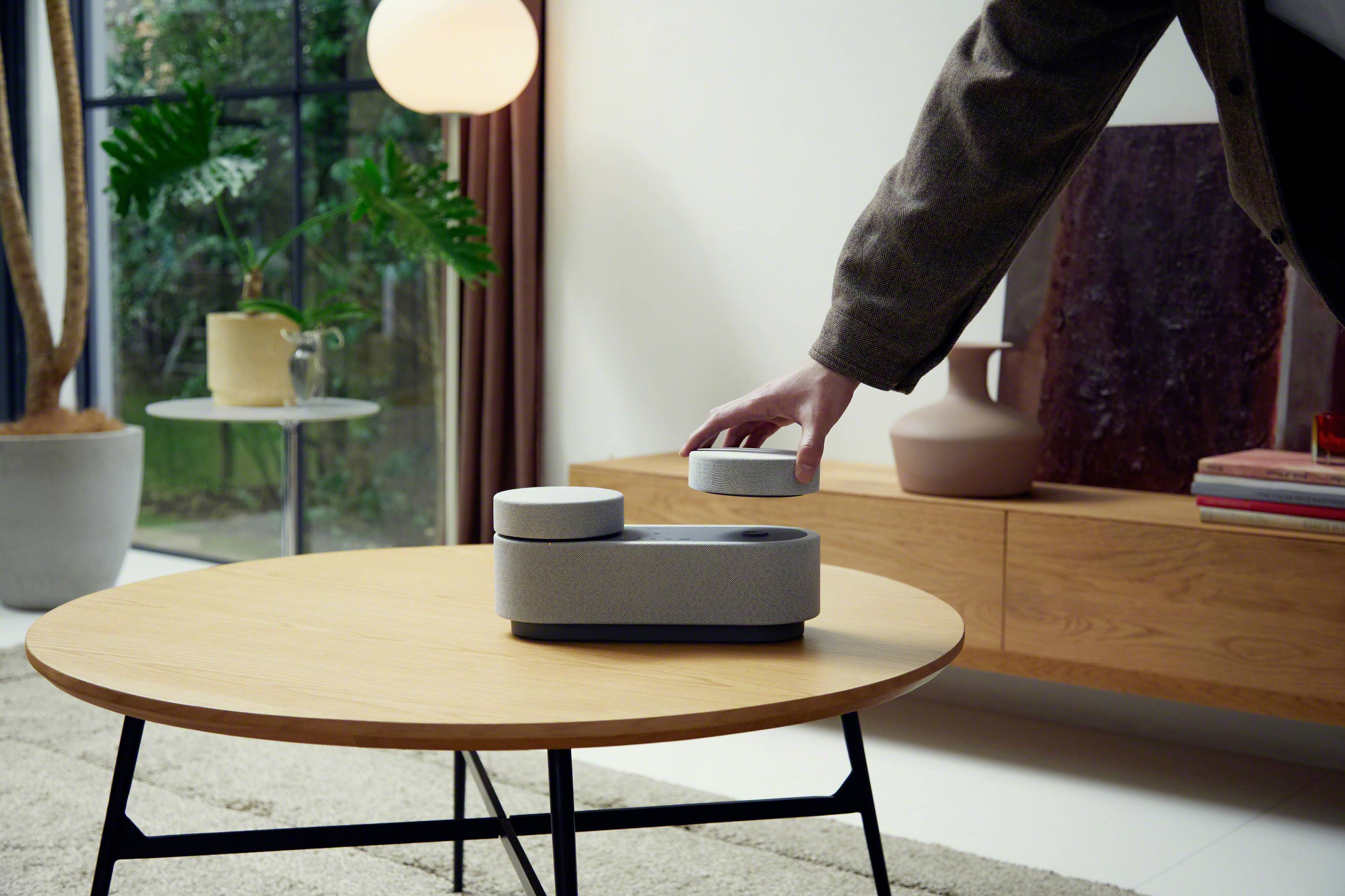 Eine Person greift ein rundes, grau-weißes Gerät auf einem Holztisch. Im Hintergrund sind Pflanzen und Möbel in einem modernen Raum zu sehen.