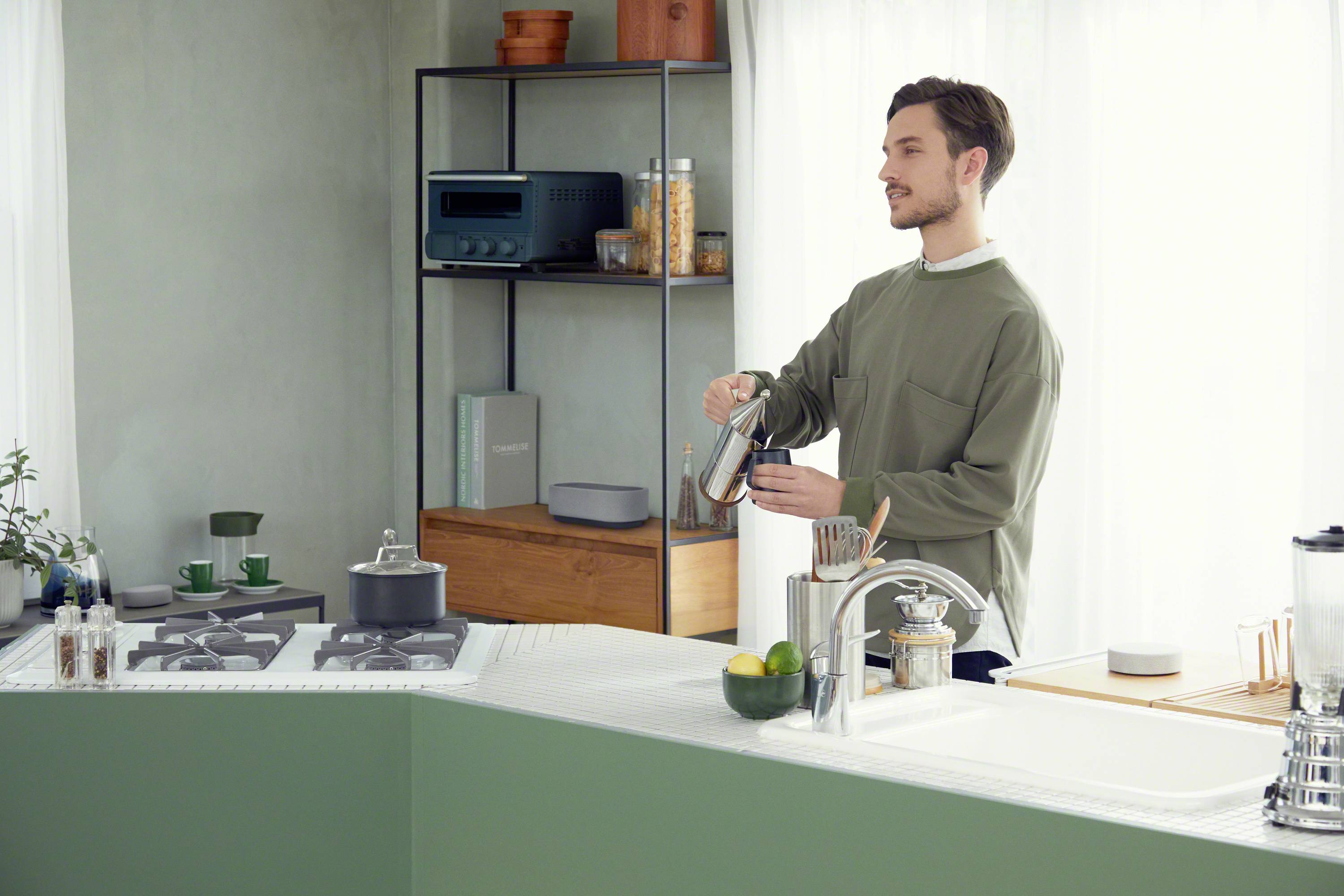 Ein Mann in einem modernen, hellen Küchenraum gießt Kaffee in eine Tasse. Im Hintergrund sind Küchenutensilien und Regale zu sehen.
