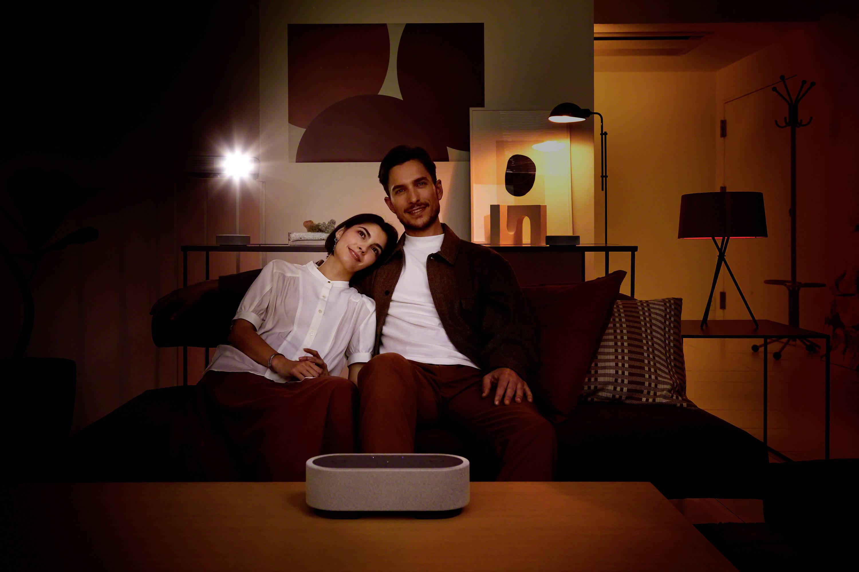 Ein Paar sitzt gemütlich auf einem Sofa in einem stimmungsvoll beleuchteten Raum. Vor ihnen steht ein grauer Lautsprecher auf dem Tisch.