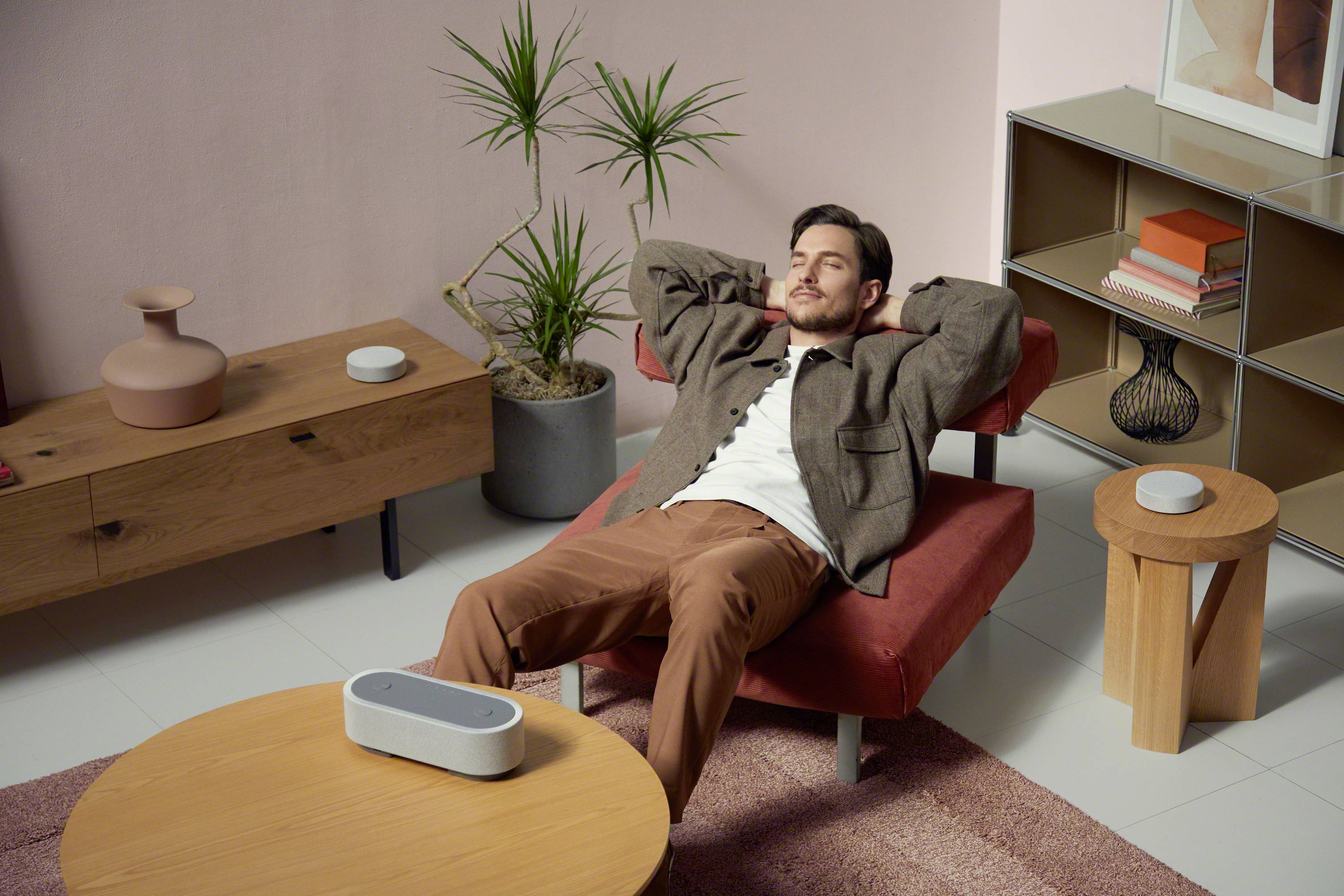 Ein Mann liegt entspannt auf einem Sessel in einem modern eingerichteten Wohnzimmer mit Pflanzen und einem Lautsprecher auf dem Tisch.