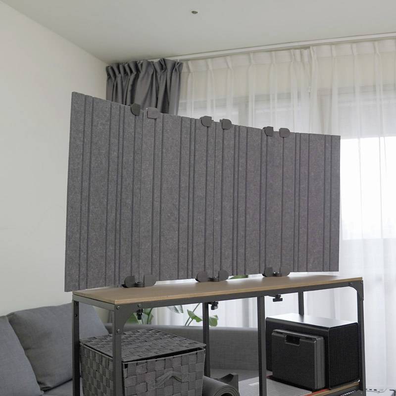 Ein Raum mit einem grauen Sofa, einem Fenster mit Vorhängen und einem Tisch mit einem akustischen Trennwand, Pflanzen und einem Lautsprecher.