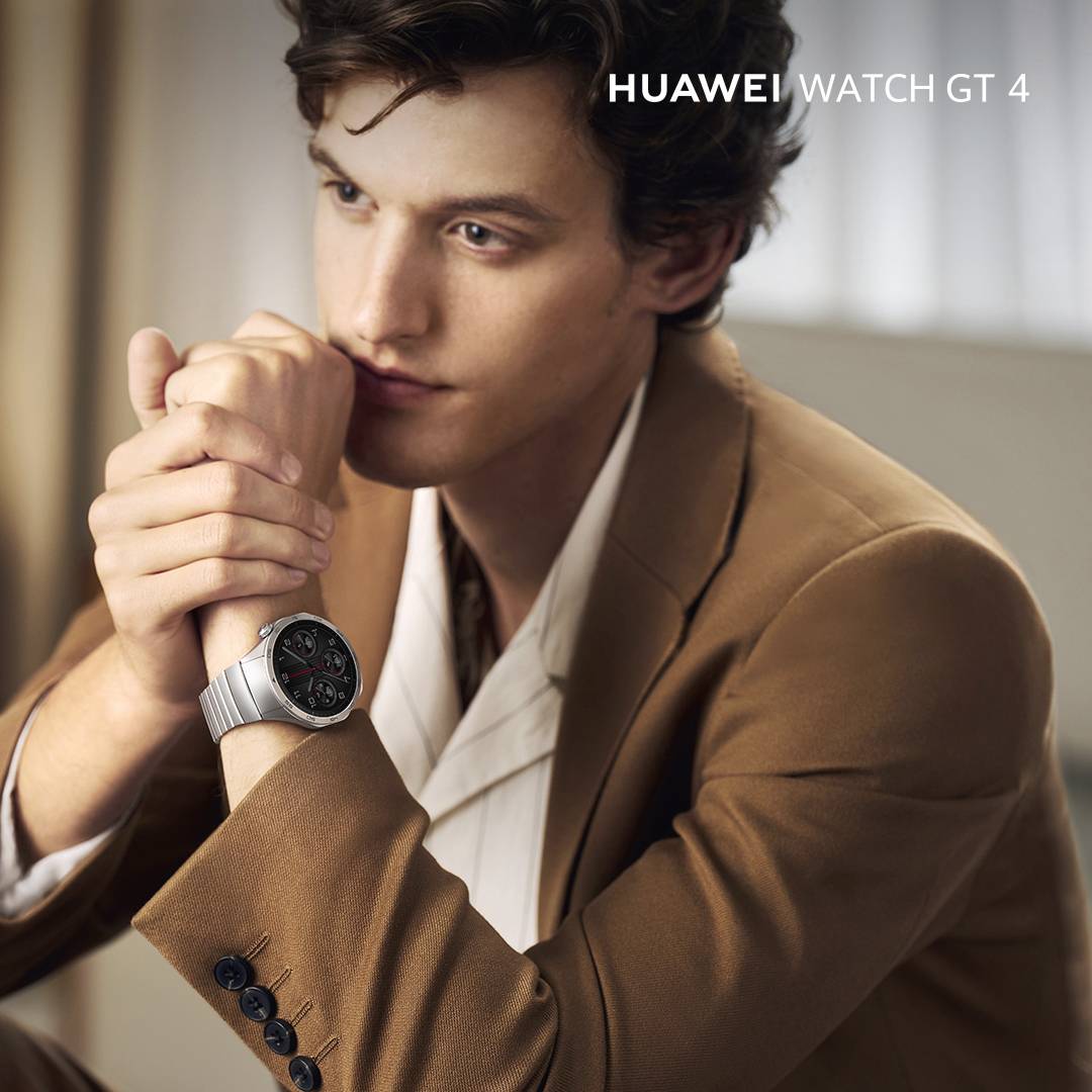 Ein Mann trägt eine 'Huawei Watch GT 4'. Die Uhr ist silber mit einem schwarzen Zifferblatt. Im Hintergrund ein diffuses Innenszenario.