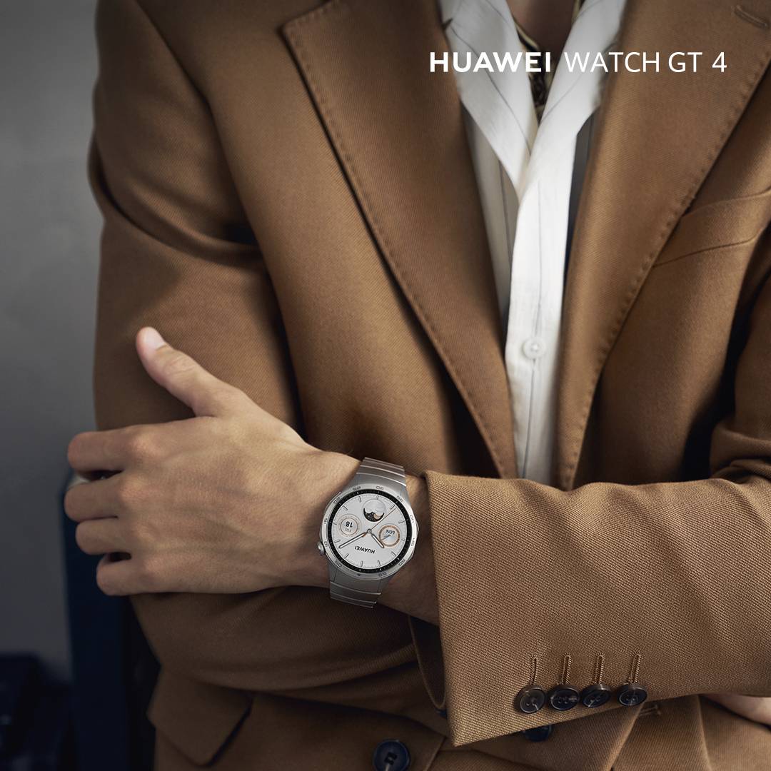 Eine Person in einem braunen Anzug trägt eine silberne Smartwatch am linken Handgelenk; die Displayanzeige ist sichtbar.