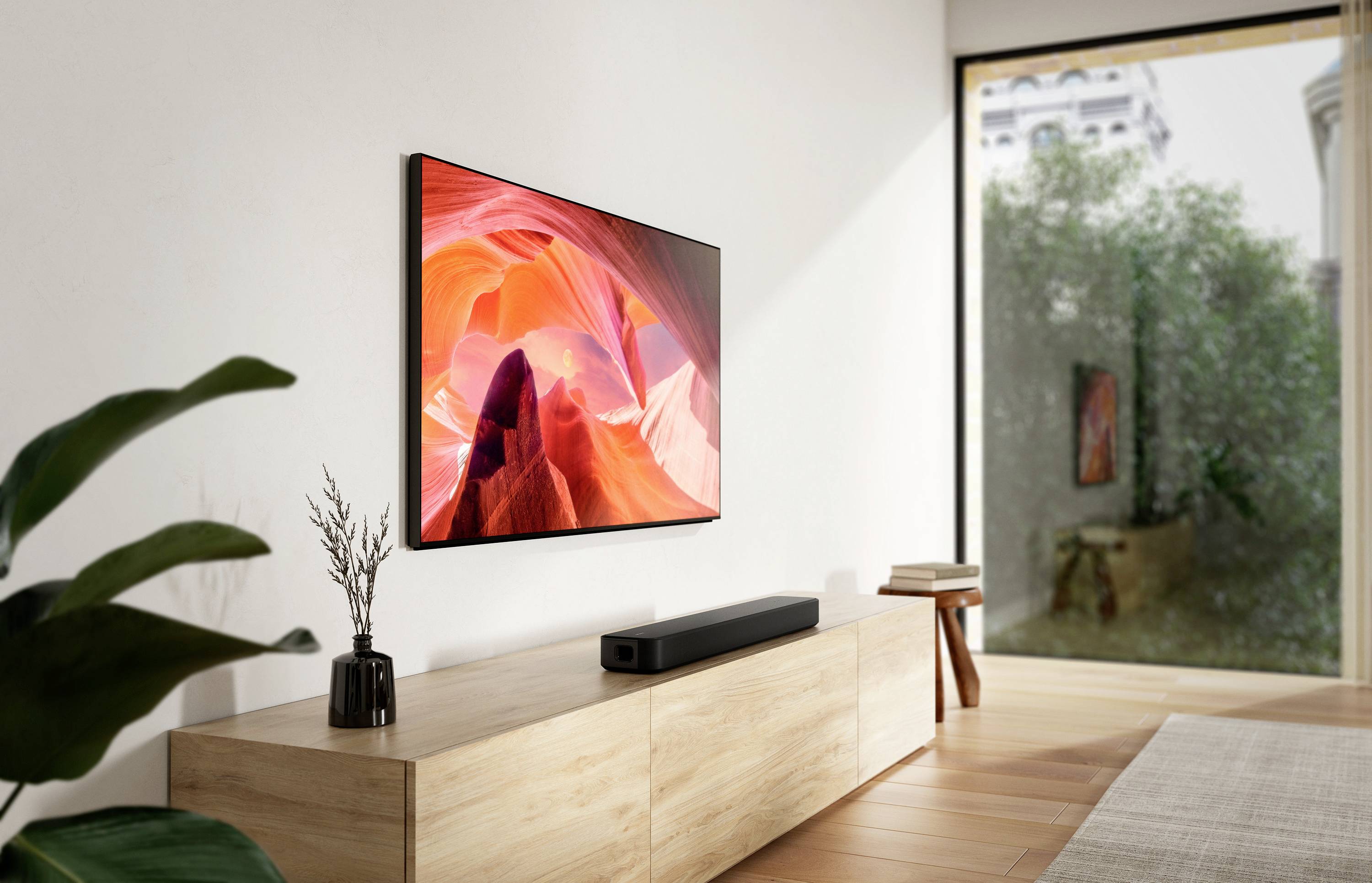 Wohnzimmer mit Holzkonsole, darauf eine Soundbar. An der Wand hängt ein großer Fernsehbildschirm, der ein Bergmotiv zeigt. Im Hintergrund ein großes Fenster mit Blick ins Grüne.