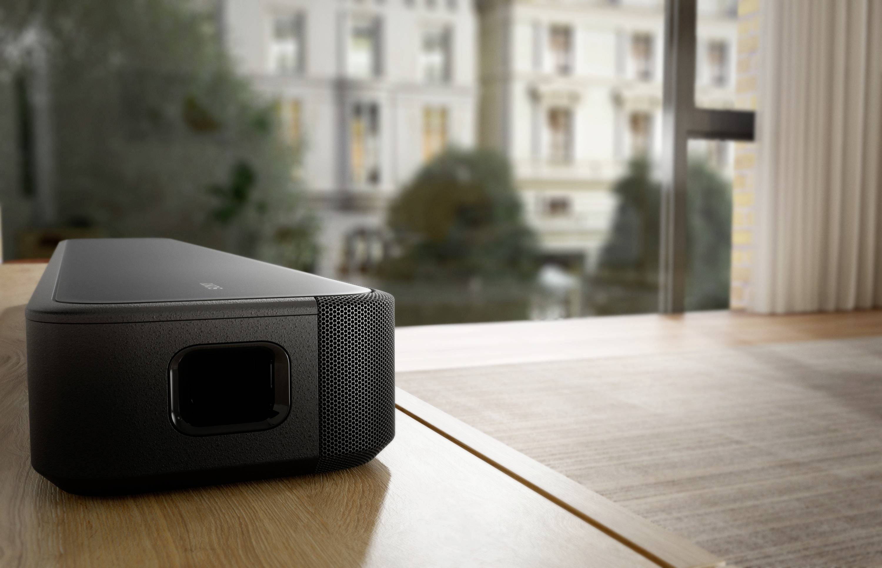 Ein schwarzer Projektor steht in einem modernen Raum auf einem Holztisch. Im Hintergrund sieht man verschwommene Gebäude durch ein Fenster.