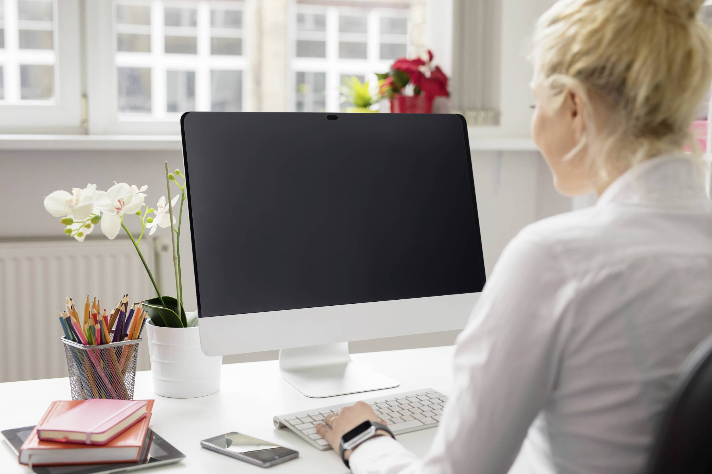 Eine Frau sitzt an einem Schreibtisch vor einem Computer mit schwarzem Bildschirm. Daneben stehen Stifte, ein Handy und Pflanzen.