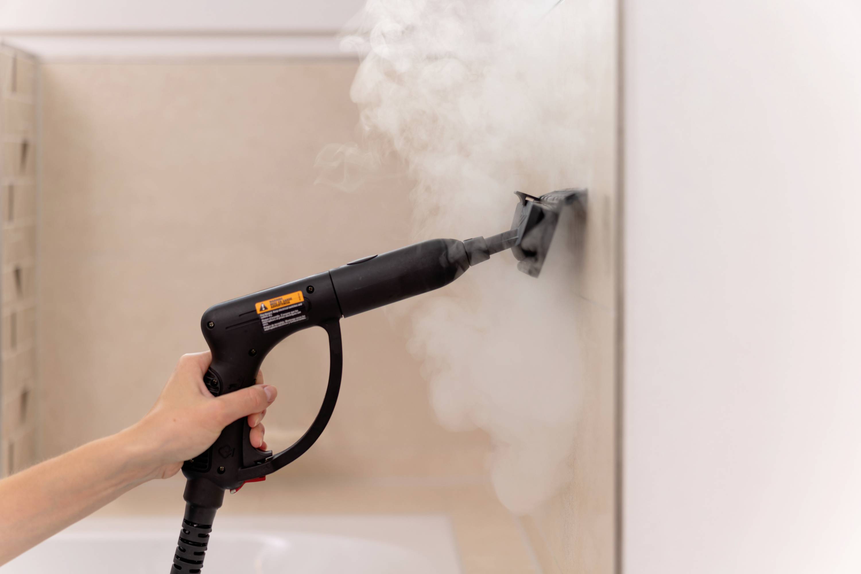 Eine Hand hält einen Dampfreiniger, der Dampf auf eine geflieste Wand sprüht, um sie zu reinigen.