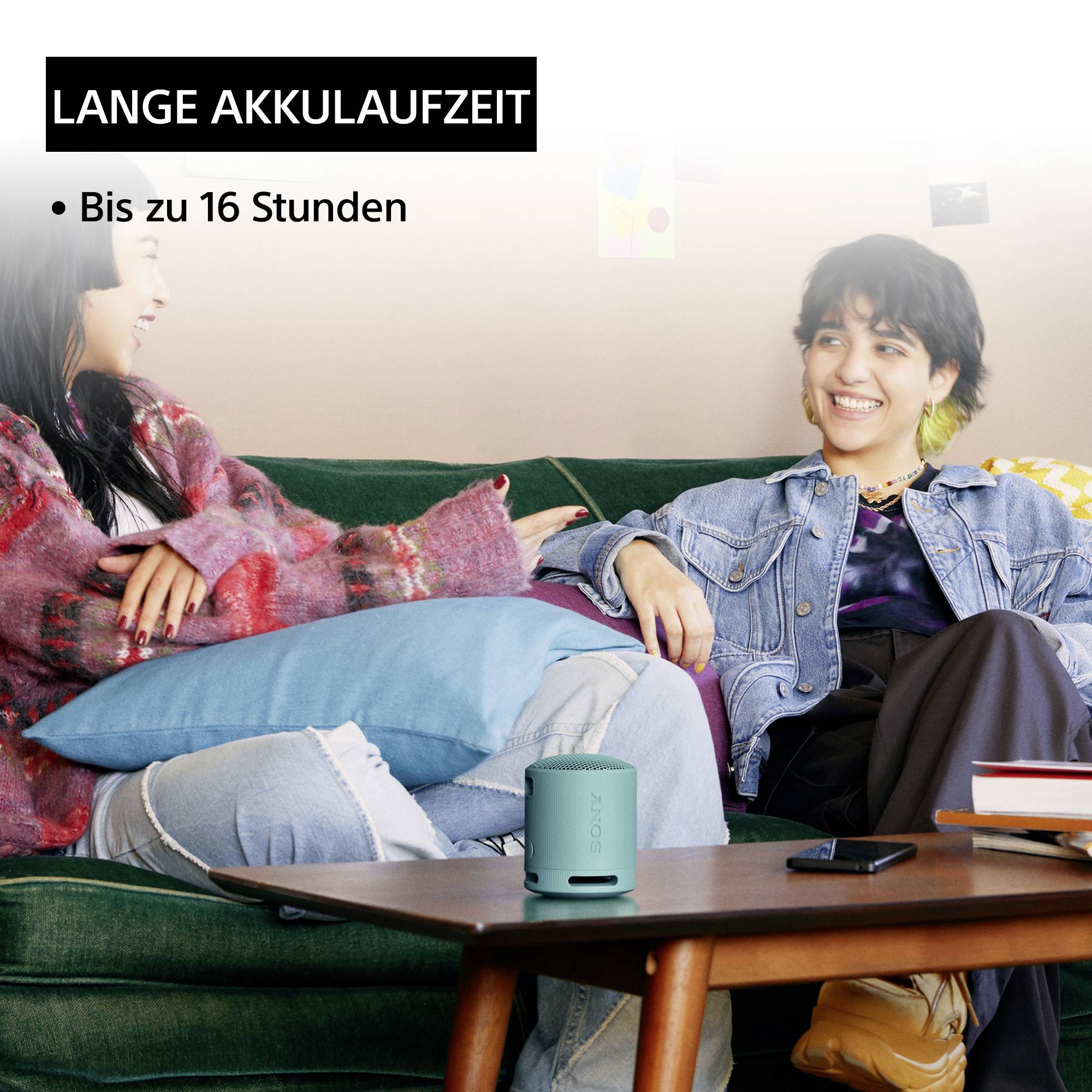 Zwei lachende Personen sitzen auf einem Sofa, eine hält einen tragbaren Lautsprecher. Text: 'Lange Akkulaufzeit. Bis zu 16 Stunden'.