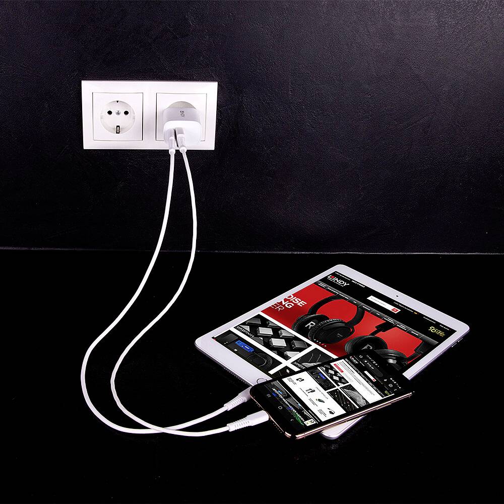 Ein Tablet und ein Smartphone werden über Ladekabel an eine Steckdose geladen. Auf dem Tablet ist eine Shopping-Website mit Kopfhörern geöffnet.