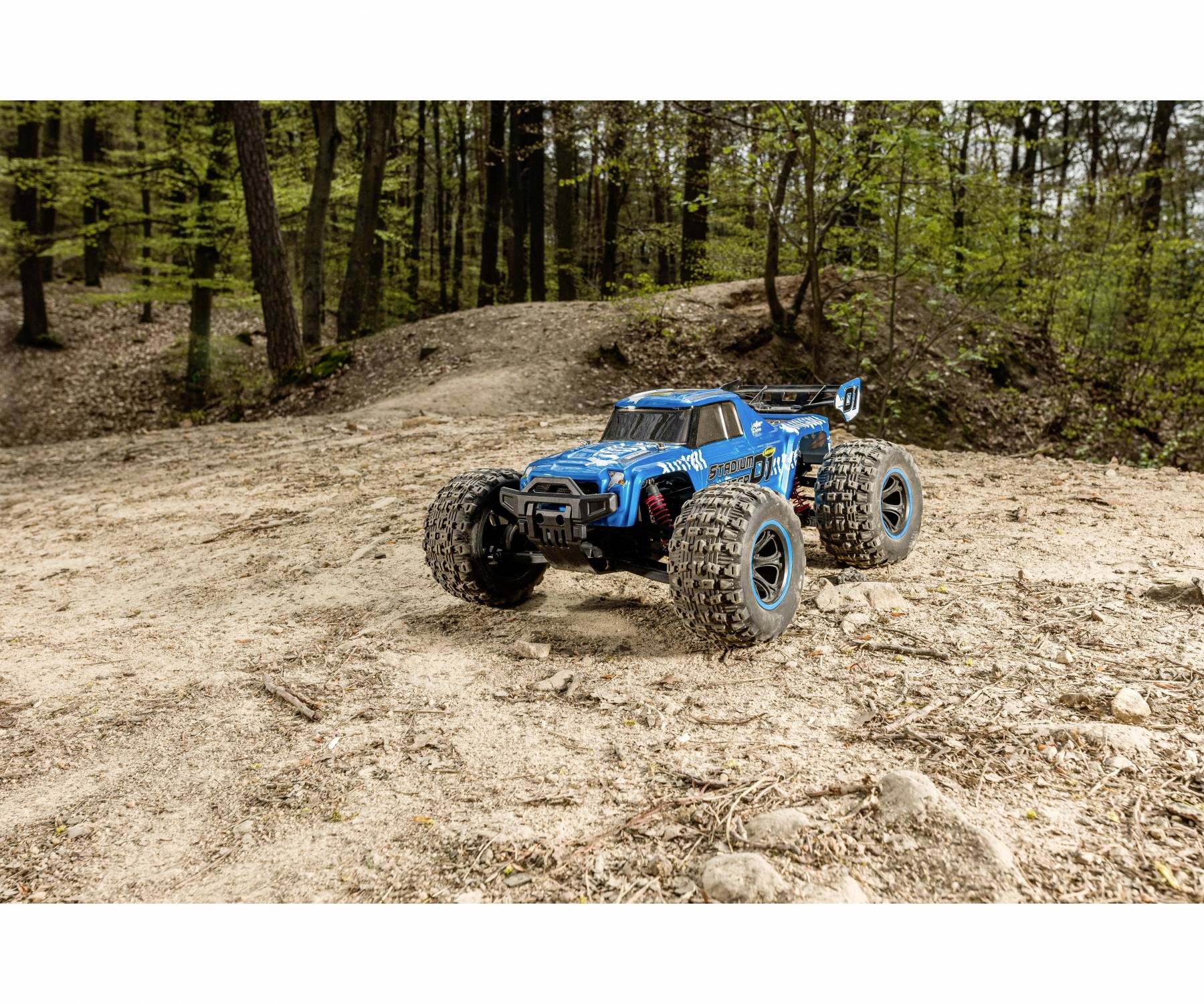Ein blaues, ferngesteuertes Auto fährt auf einem sandigen Weg im Wald. Bäume umgeben die Szene, während das Auto im Vordergrund steht.
