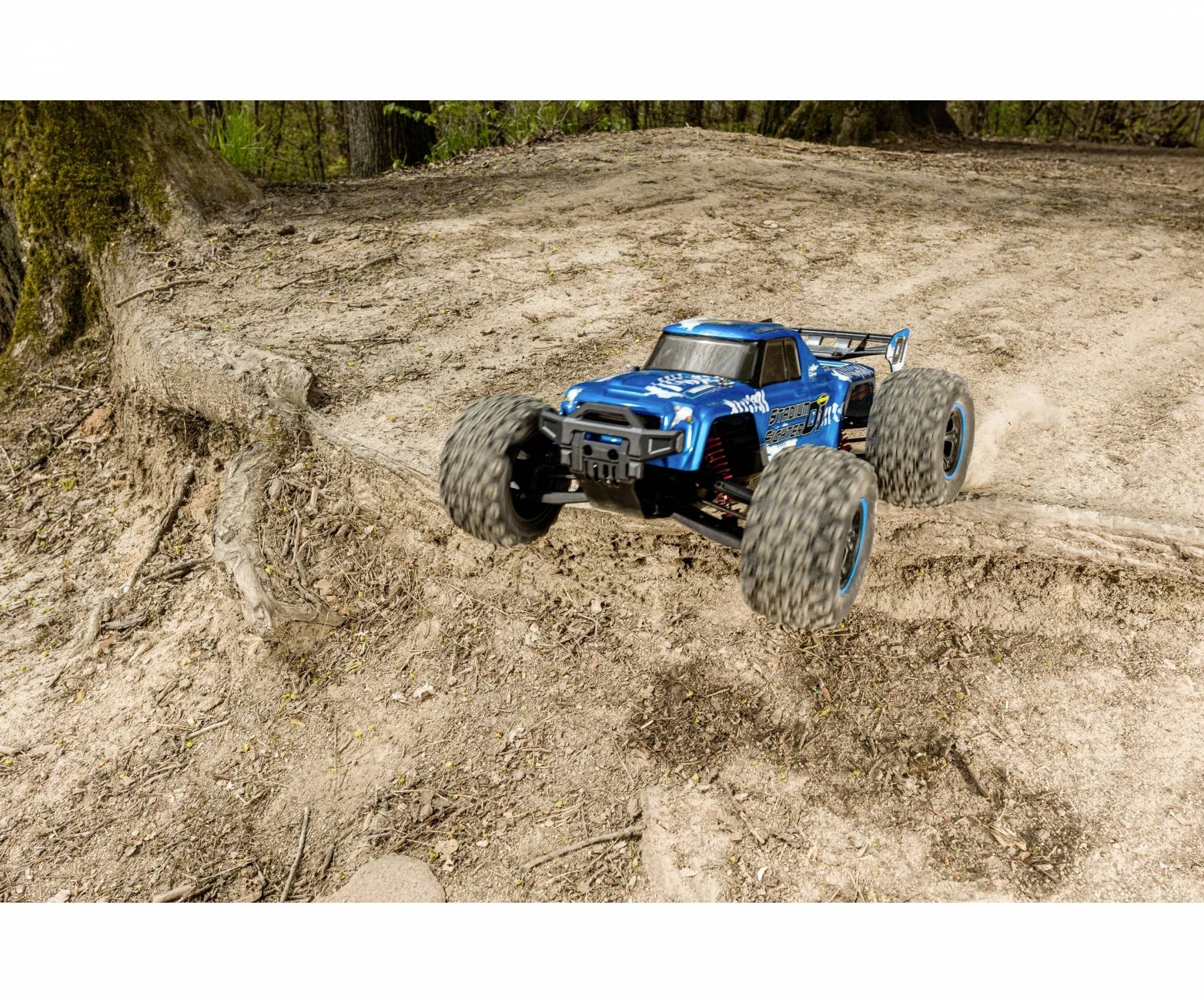 Ein blauer ferngesteuerter Geländewagen springt über einen sandigen Hügel in einem Waldgebiet.