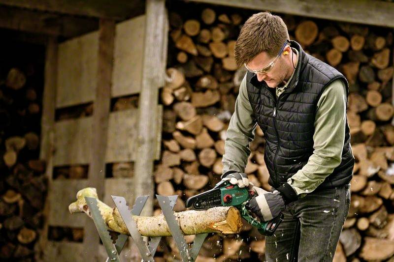 Ein Mann mit Sicherheitsbrille und Gehörschutz zersägt mit einer Motorsäge einen Holzstamm auf einer Sägebockvorrichtung. Im Hintergrund ist gestapeltes Brennholz zu sehen.