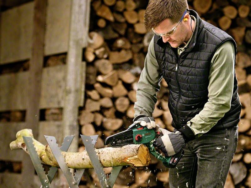 Mann sägt Holzscheit mit einer elektrischen Kettensäge in Werkstatt. Stapel von Brennholz im Hintergrund.