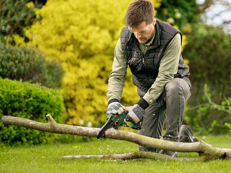 Ein Mann mit Schutzbrille und Handschuhen kniet im Garten und sägt mit einer Motorsäge einen Baumstamm. Im Hintergrund sind Büsche.
