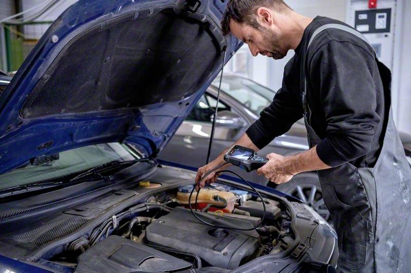 Ein Mechaniker inspiziert den Motor eines Autos mit einem Diagnosegerät in einer Werkstatt. Im Hintergrund steht ein weiteres Auto.