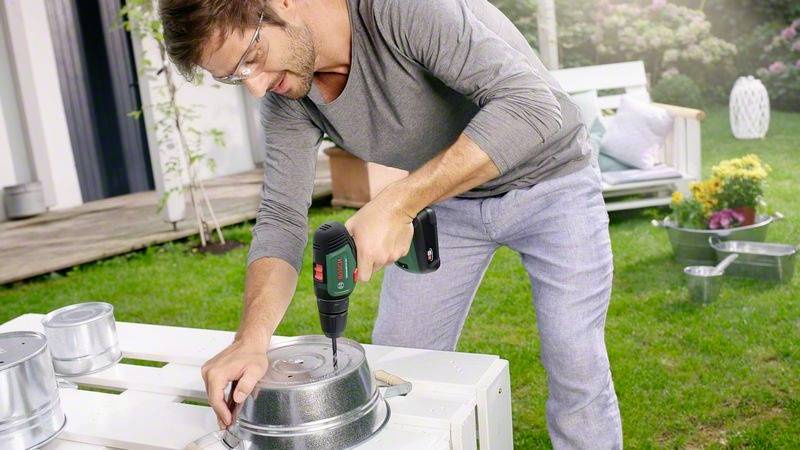 Ein Mann in Freizeitkleidung bohrt mit einem Akkuschrauber ein Loch in einen Metalleimer auf einer Terrasse im Garten.
