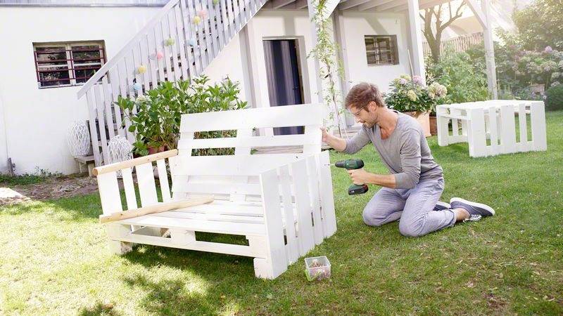 Eine Person baut im Garten eine weiße Holzbank zusammen. Im Hintergrund ist ein Haus mit Terrasse sichtbar.