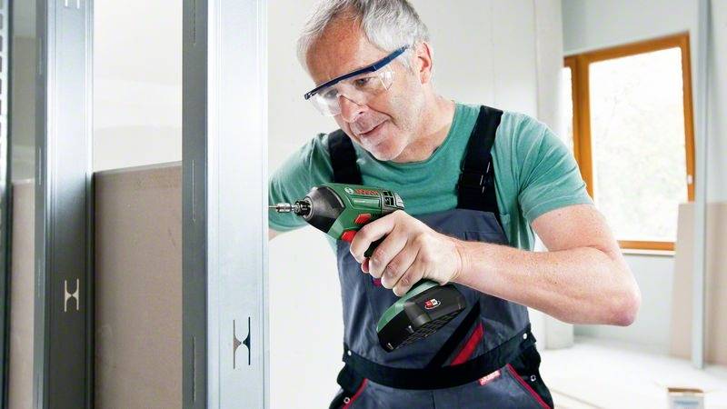 Ein älterer Mann mit Schutzbrille und grüner Latzhose verwendet einen Akkuschrauber, um Metallrahmen in einem hellen Raum zu montieren.