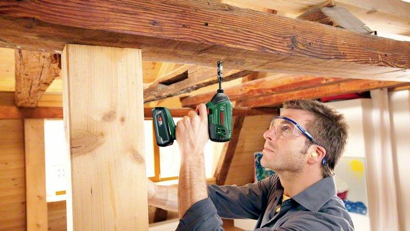 Ein Mann bohrt mit einem Akkuschrauber in einen Holzbalken unter einer Holzdecke. Er trägt Schutzbrille und Gehörschutz.