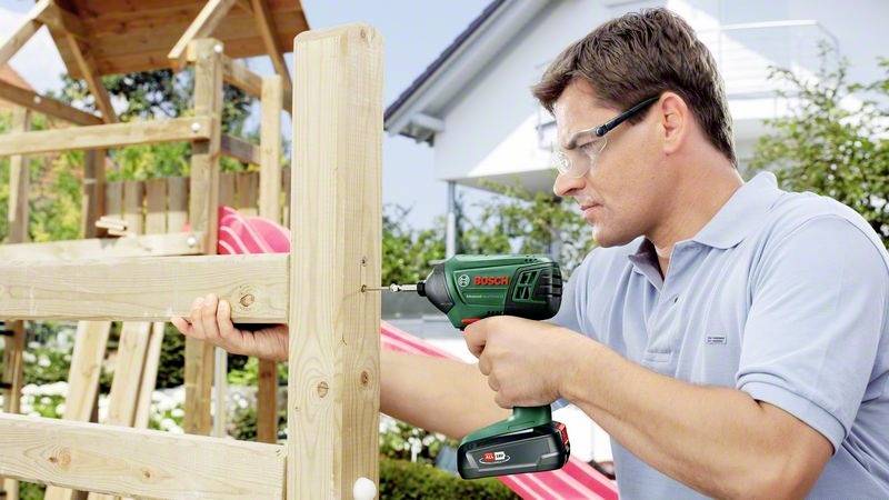 Ein Mann in Schutzbrille verwendet einen Akkuschrauber, um ein Holzgerüst im Freien zu befestigen. Im Hintergrund steht ein Haus.