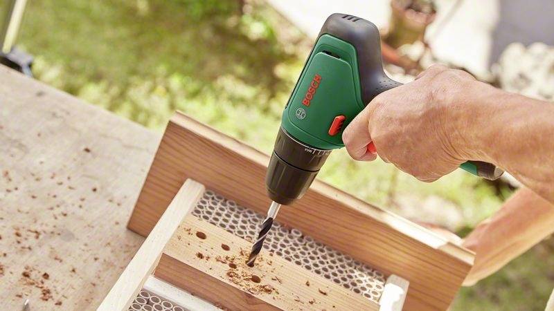 Eine Hand hält einen Bohrer, der ein Holzbrett auf einem Tisch durchbohrt. Holzspäne sind um das Bohrloch verteilt.