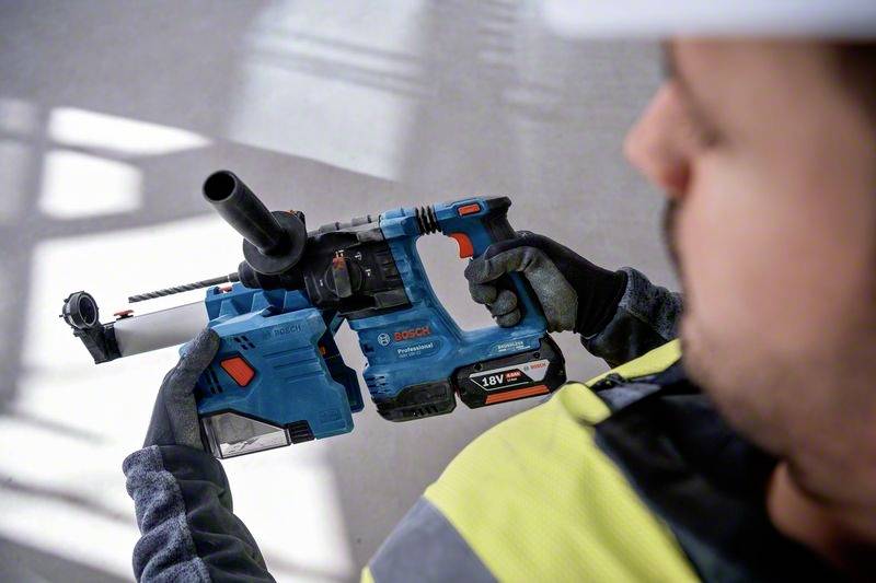 Ein Arbeiter hält einen blauen Bohrhammer mit Staubabsaugung, gekleidet in Schutzkleidung. Hintergrund unscharf, Fokus auf das Werkzeug.