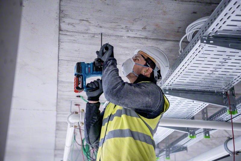Ein Arbeiter in Schutzkleidung und Helm bohrt in eine Decke, umgeben von Kabeln und Rohren. Er trägt eine Schutzmaske.