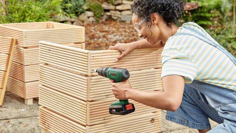 Eine Person mit Schutzbrille verwendet eine Bohrmaschine, um einen Holzkasten im Freien zusammenzubauen. Pflanzen und Steine im Hintergrund.