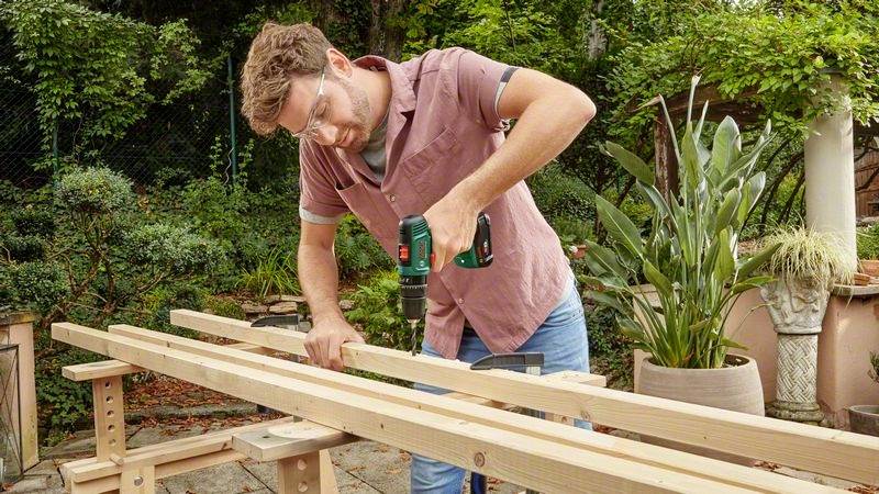 Ein Mann bohrt im Freien ein Loch in ein Holzbrett. Er trägt eine Schutzbrille und verwendet eine grüne Bohrmaschine. Im Hintergrund sind Pflanzen sichtbar.
