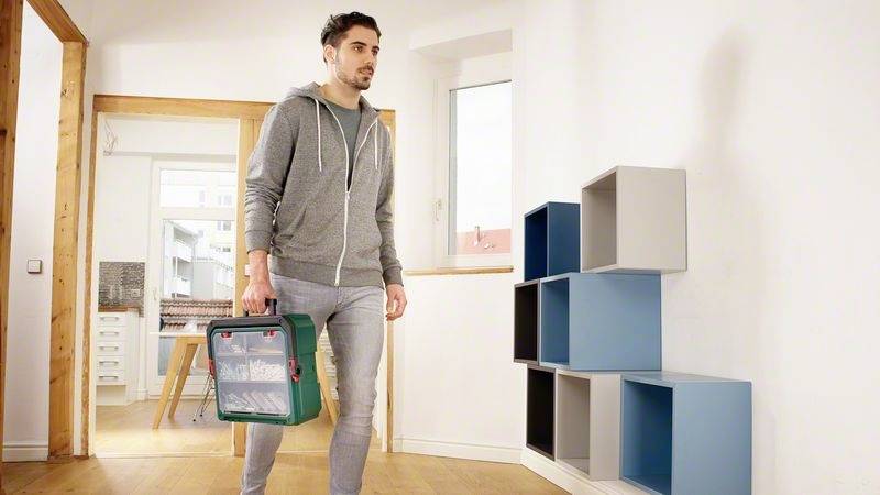 Ein Mann in Freizeitkleidung trägt eine Werkzeugkiste durch einen Raum mit bunten Wandregalen.
