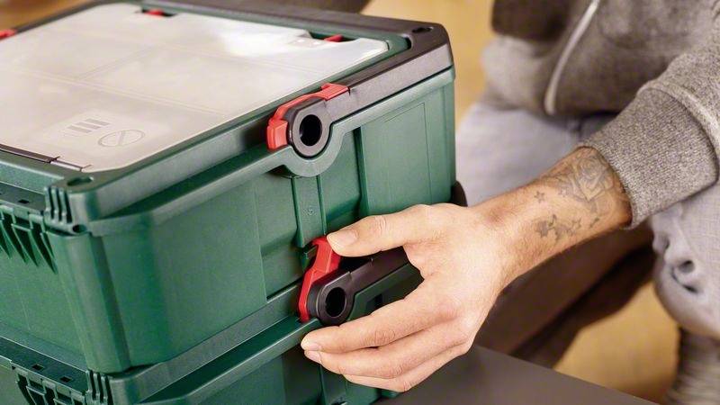 Eine Person stapelt zwei grüne Werkzeugboxen, indem sie rote Clips an den Seiten verriegelt.
