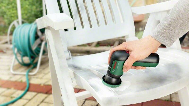 Eine Person reinigt einen weißen Plastikstuhl mit einem kleinen, handgehaltenen Reinigungsgerät. Im Hintergrund ist ein Gartenschlauch zu sehen.