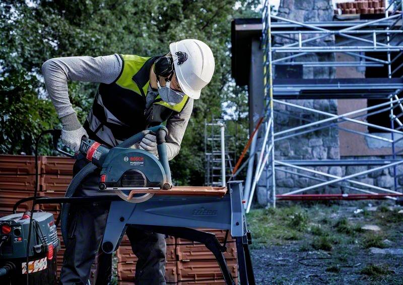 Arbeiter mit Schutzhelm und Maske schneidet mit Elektrosäge Holz auf einer Baustelle, im Hintergrund steht ein Gerüst.