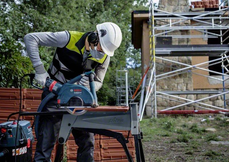 Ein Bauarbeiter mit Schutzhelm und Sicherheitskleidung verwendet ein elektrisches Werkzeug, um Rohre auf einer Baustelle zu schneiden. Im Hintergrund ist ein Gerüst am Gebäude sichtbar.