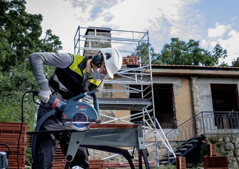 Ein Bauarbeiter mit Schutzhelm und Gehörschutz benutzt eine Kreissäge auf einer Baustelle. Im Hintergrund ist ein Gerüst sichtbar.