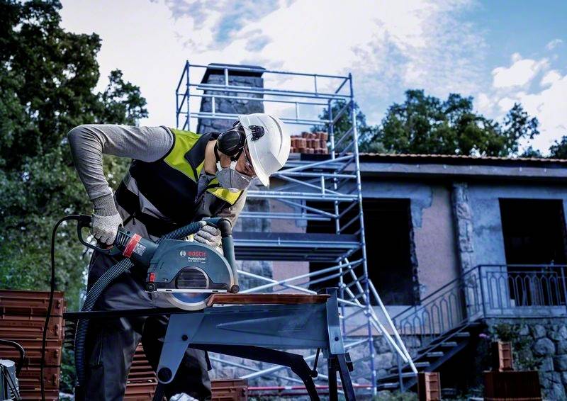 Ein Bauarbeiter mit Schutzhelm und Gehörschutz verwendet eine Kreissäge, um ein Metallstück vor einem Gerüst und Gebäude zu schneiden.
