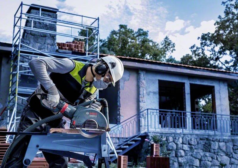 Ein Bauarbeiter mit Schutzkleidung bedient eine elektrische Säge vor einem im Bau befindlichen Gebäude mit Gerüst im Hintergrund.