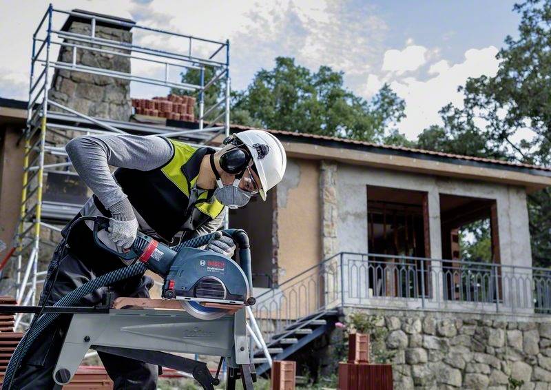 Ein Bauarbeiter mit Schutzhelm und Gehörschutz schneidet Holz mit einer elektrischen Säge auf einer Baustelle vor einem Gebäude.