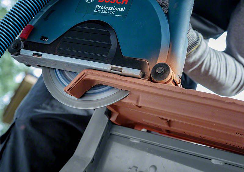 Eine Hand hält eine Elektrosäge, die gerade eine Dachziegel schneidet. Staub wird von einem Sauger abgesaugt.